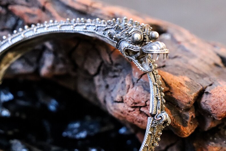 Pu&ograve; includere: Braccialetto d'argento con un designo dettagliato e ornato che presenta una testa di creatura mitologica con un becco e denti affilati. Il braccialetto &egrave; aperto e poggia su un pezzo di legno invecchiato.