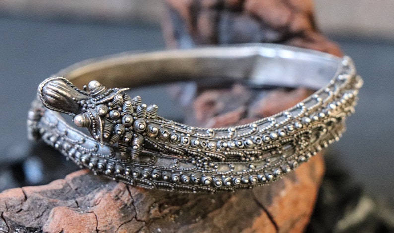 Pu&ograve; includere: Un braccialetto d'argento con dettagli intricati, che presenta una testa di drago con un motivo a perline.