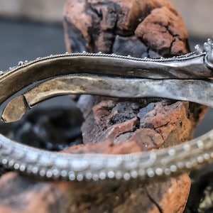 Pu&ograve; includere: Braccialetto d'argento con dettagli intricati e una chiusura a testa di drago. Il braccialetto &egrave; aperto e appoggiato su un pezzo di legno.
