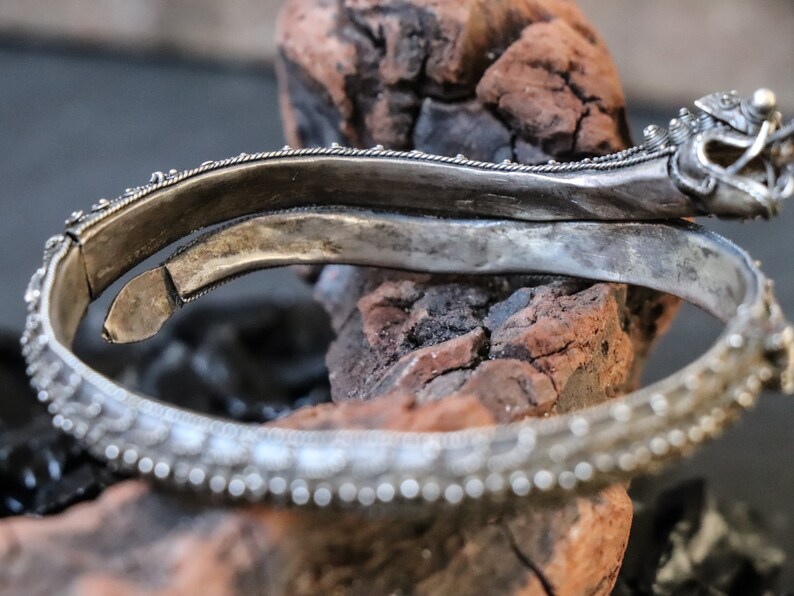 Pu&ograve; includere: Braccialetto d'argento con dettagli intricati e una chiusura a testa di drago. Il braccialetto &egrave; aperto e appoggiato su un pezzo di legno.