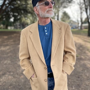 May include: A man wearing a tan blazer, a blue shirt, and blue jeans. He is wearing a black cap and sunglasses.