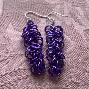 Może przedstawiać: Para fioletowych kolczyków z łańcuszka. Każdy kolczyk składa się z szeregu małych, połączonych pierścieni, tworzących teksturowany, zwisający wzór. Kolczyki mają srebrne haczyki. Tło to teksturowana, jasnofioletowa tkanina.