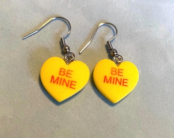 May include: A pair of yellow heart-shaped earrings with the words "BE MINE" in red lettering. The earrings are on a white background.
