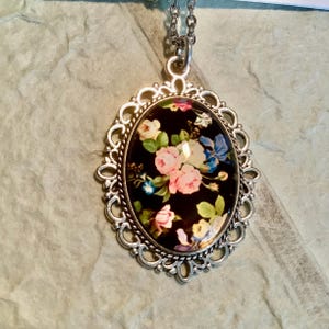 May include: A silver oval pendant necklace with a black background and a floral design featuring pink, white, and blue flowers. The pendant has a decorative filigree border.