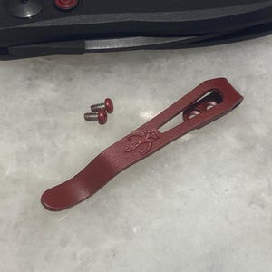 May include: A red metal pocket clip with a small, engraved design. The clip has three holes and is designed to attach to a pocket knife.