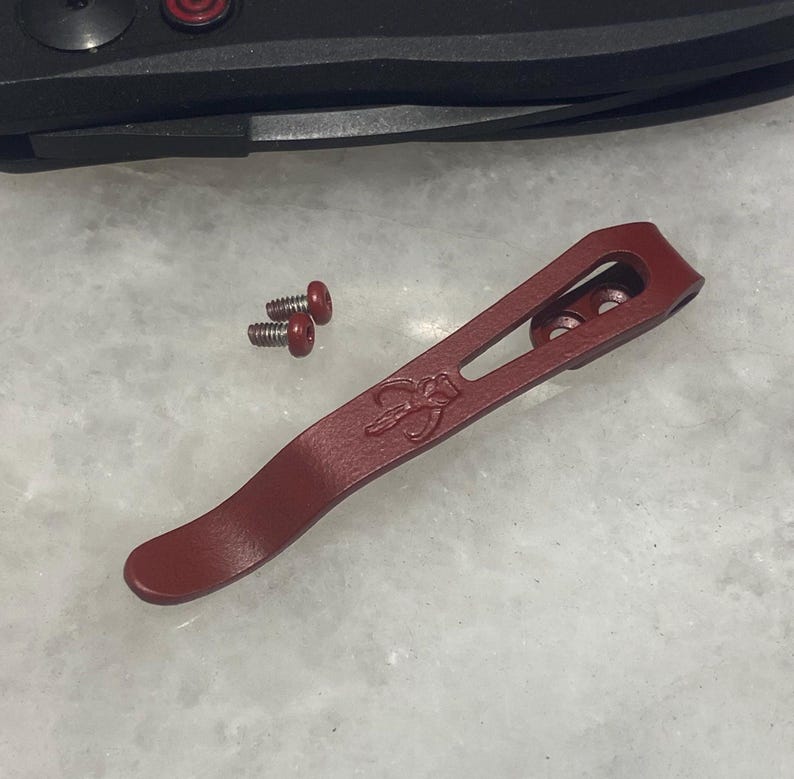 May include: A red metal pocket clip with a small, engraved design. The clip has three holes and is designed to attach to a pocket knife.