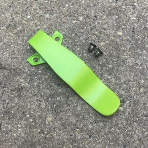 May include: A bright green pocket clip with two mounting holes and two small screws. The clip is a vibrant, solid color and has a smooth, curved design. The screws are dark metal and appear to be new.