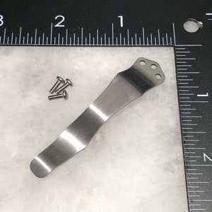 May include: A silver metal pocket clip with three holes at the top. There are four small silver screws next to the clip.
