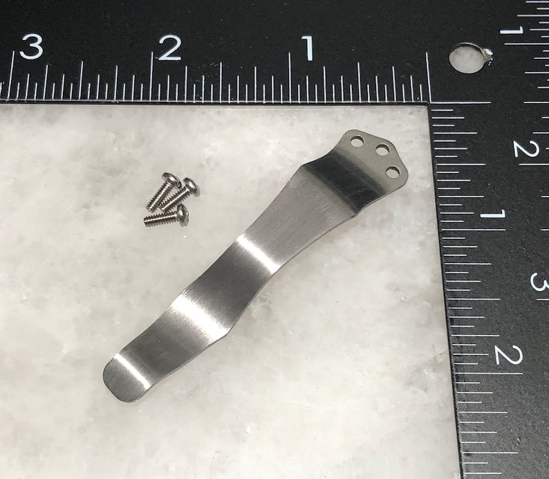 May include: A silver metal pocket clip with three holes at the top. There are four small silver screws next to the clip.