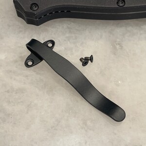 Puede incluir: Clip de bolsillo de metal negro con dos pequeños tornillos. El clip es curvo y tiene un acabado liso.