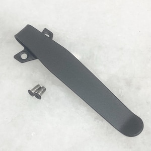 May include: A matte black pocket clip with two small silver screws. The clip is long and curved, with a rounded end and two holes for attachment. The clip is designed to attach to a knife or other item.
