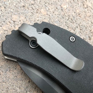 May include: A black pocket knife with a titanium pocket clip. The knife has a textured black handle and a partially visible blade.