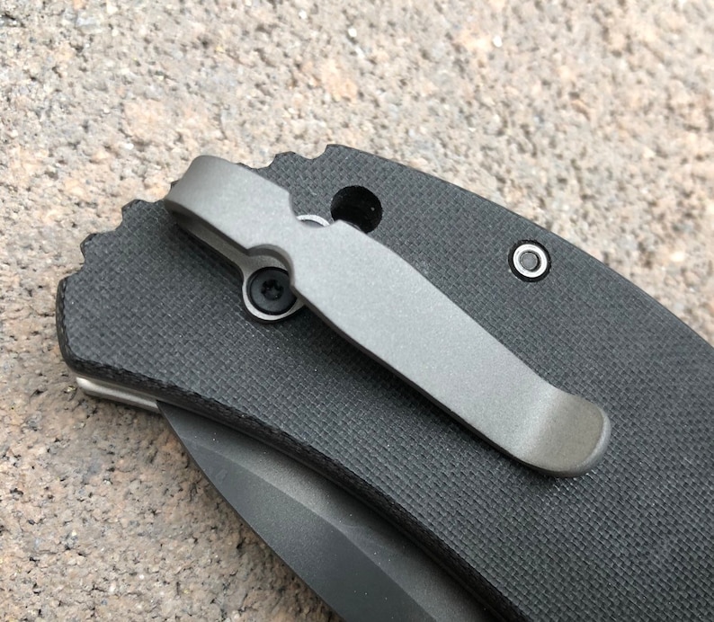 May include: A black pocket knife with a titanium pocket clip. The knife has a textured black handle and a partially visible blade.