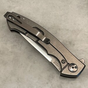 May include: A silver pocket knife with a partially open blade. The knife has a pocket clip and a textured handle. The blade is partially open and has a sharp point. The knife has the text "0650 EAB" and "KNIFE USA" on the handle.