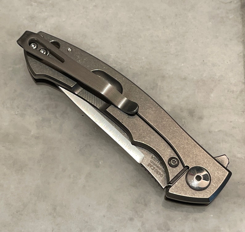 May include: A silver pocket knife with a partially open blade. The knife has a pocket clip and a textured handle. The blade is partially open and has a sharp point. The knife has the text "0650 EAB" and "KNIFE USA" on the handle.