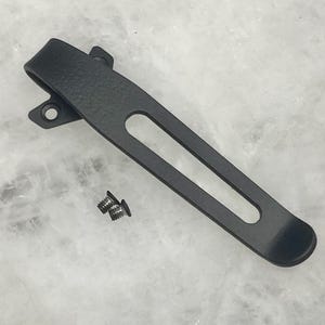 May include: A black pocket clip with a textured finish and a long, oval-shaped opening. Two small screws are included. The clip has two holes for mounting. The clip is designed for attaching to a pocket or other item.