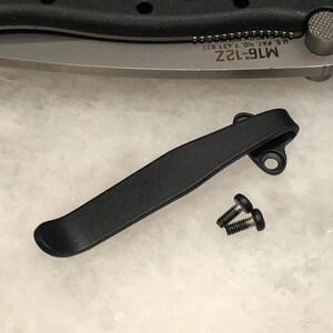 May include: Black metal pocket clip and two small screws. The clip is curved and has two holes for mounting. The knife blade is visible in the background, with the text "M16-12Z" and "U.S. PAT. NO. 7,437,022".