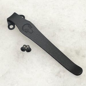 May include: Black metal pocket clip with a skull design and two screws.