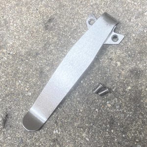 May include: A silver-colored metal pocket clip with a brushed finish, featuring a wide mounting base with two pre-drilled circular holes for attachment. The clip is accompanied by two small, threaded metal screws for secure installation.