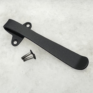 May include: A black metal pocket clip with two screw fasteners. The clip has a curved design with two holes for mounting. The fasteners are silver in color.