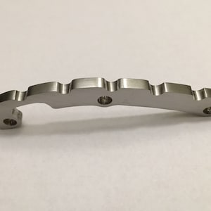 May include: A silver metal part with a curved shape and five holes. The part has a textured surface and appears to be a component for a mechanical device.