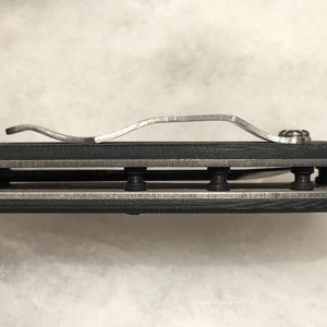 May include: Close-up of a black folding knife with a silver clip and a silver blade. The knife is closed and the clip is attached to the handle.
