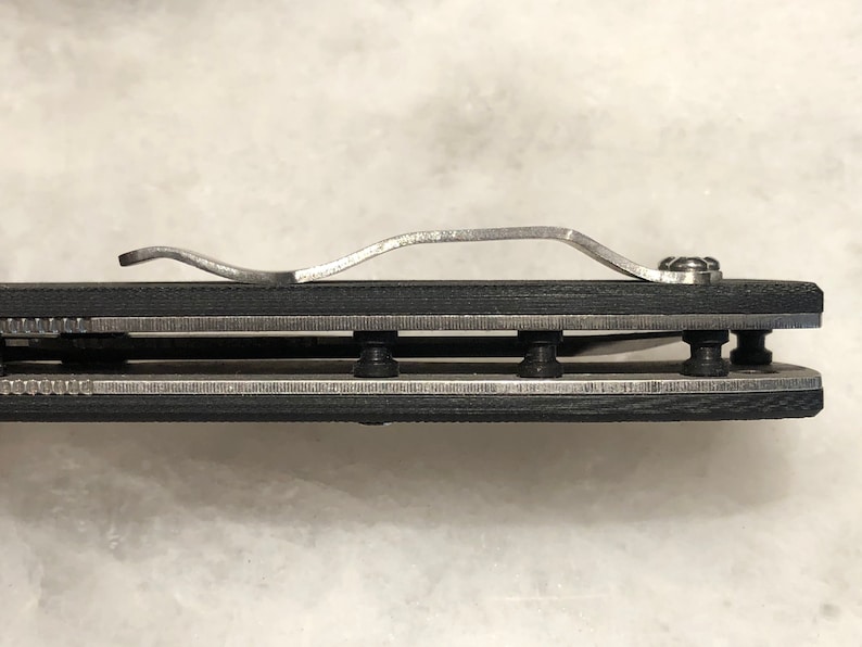 May include: Close-up of a black folding knife with a silver clip and a silver blade. The knife is closed and the clip is attached to the handle.