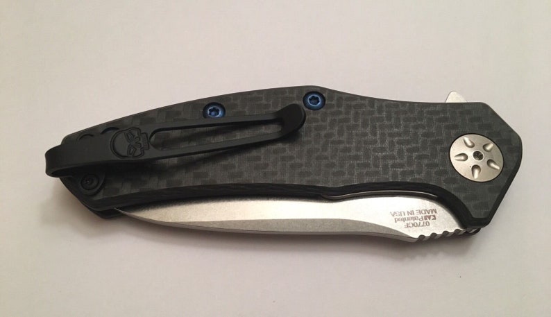 Puede incluir: Un cuchillo de bolsillo negro con un mango de fibra de carbono y una hoja plateada. El cuchillo tiene un logotipo de calavera en el clip y un bot&oacute;n plateado en la parte posterior. La hoja est&aacute; parcialmente abierta y el texto "GT20CF" y "EAR-PENNED MADE IN USA" son visibles en la hoja.