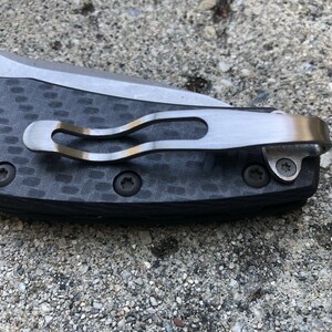 May include: A close-up of a black carbon fiber pocket knife with a silver clip and two screws. The knife is made in the USA.