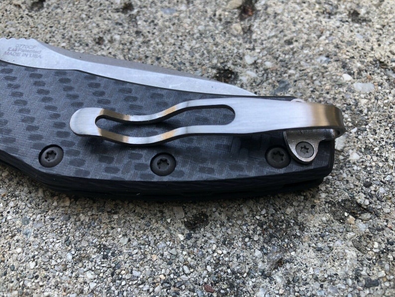 May include: A close-up of a black carbon fiber pocket knife with a silver clip and two screws. The knife is made in the USA.