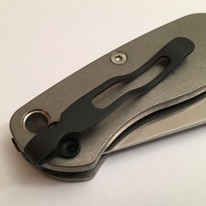 May include: A close-up of a silver pocket knife with a black pocket clip. The knife is closed and the blade is not visible.