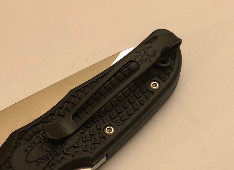 May include: Black pocket knife with a textured black handle and a black pocket clip. The pocket clip has a spider logo on it.