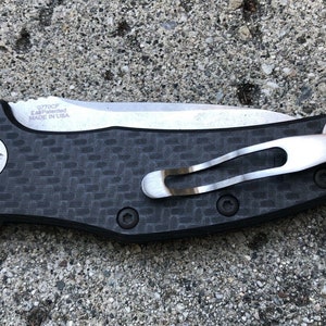 May include: A black and silver pocket knife with a carbon fiber handle and a deep carry pocket clip. The knife has a  "ZT 0770CF EAF Patented Made in USA" inscription on the blade.