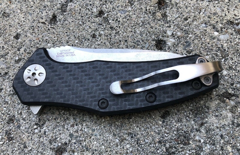 May include: A black and silver pocket knife with a carbon fiber handle and a deep carry pocket clip. The knife has a  "ZT 0770CF EAF Patented Made in USA" inscription on the blade.