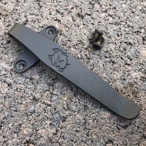 May include: Black metal pocket clip with a wolf head logo. The clip is designed to attach to a pocket knife or other tools. Two screws are included.