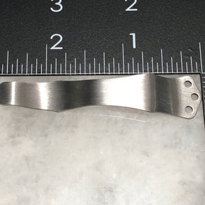 May include: A silver metal pocket clip with three holes. The clip is curved and designed to attach to a pocket.