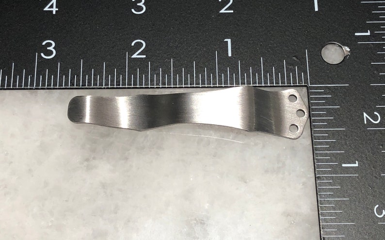 May include: A silver metal pocket clip with three holes. The clip is curved and designed to attach to a pocket.
