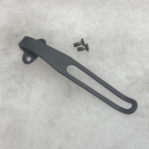 May include: A black metal pocket clip for a knife. The clip has a simple design with a single loop and a small hole at the top. Two small screws are included.