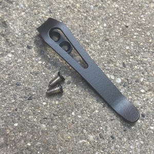 May include: A matte black metal pocket clip with a curved top and an elongated cutout, accompanied by two small silver-toned metal screws with black heads, designed for attachment to a folding tool or knife handle for secure carry.