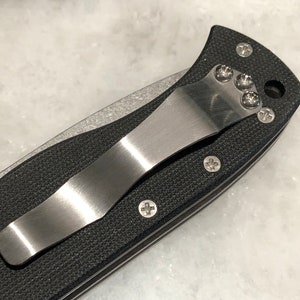 May include: Close-up of a black pocket knife with a stainless steel clip. The clip is attached to the knife with four screws.