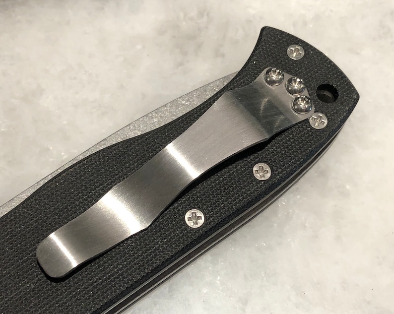May include: Close-up of a black pocket knife with a stainless steel clip. The clip is attached to the knife with four screws.