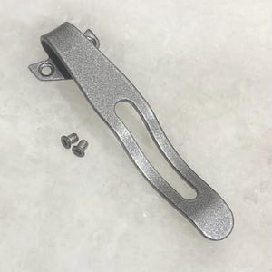 May include: A silver-colored metal pocket clip with two small screws. The clip has a curved design with a cutout in the center and a mounting bracket with two holes. The clip is likely for attaching to a knife or other tool.