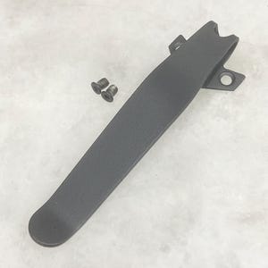 May include: A dark gray pocket clip for a knife, with a textured surface, and two small screws. The clip has a curved design with a rounded end and a notched top, and two holes for mounting.