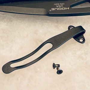 May include: Black metal pocket clip with two screws. The clip is designed to attach to a pocket knife or other tool. The clip is made of a durable metal and has a smooth finish.