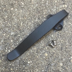May include: A matte black metal pocket clip with a curved, elongated design featuring a triangular mounting base with two circular screw holes, accompanied by two small silver-coloured flat-head screws for attachment purposes.