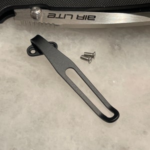 May include: Black metal pocket clip with two silver screws. The clip is designed to attach to a pocket knife or other tool.
