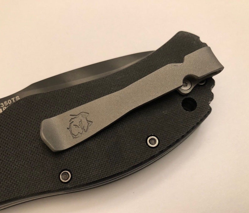 Puede incluir: Un cuchillo de bolsillo negro con un clip de bolsillo de titanio. El clip tiene un logotipo grabado.