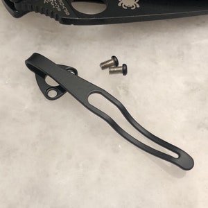 May include: A black metal pocket clip with two small screws. The clip has a curved design with a loop and two mounting holes. The screws are silver with black heads. The items appear to be for a knife.