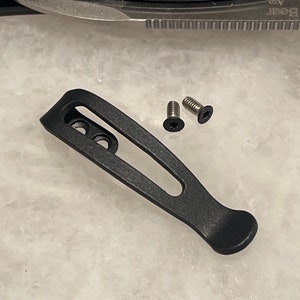 May include: Black metal pocket clip with three screw holes and two screws. The clip is designed to attach to a pocket or belt.