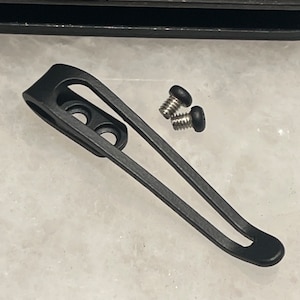 Może przedstawiać: Czarny metalowy klips do kieszeni z dwoma małymi śrubami. Klips jest zaprojektowany do mocowania do kieszeni lub paska.